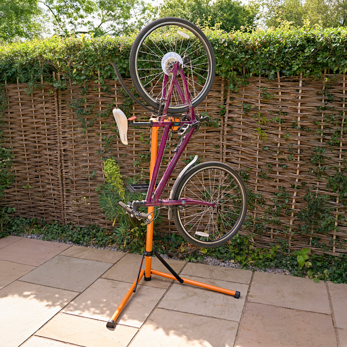 Soporte de reparación para bicicletas en color naranja y negro, sostiene una bicicleta de montaña de color púrpura al revés sobre un fondo de seto verde