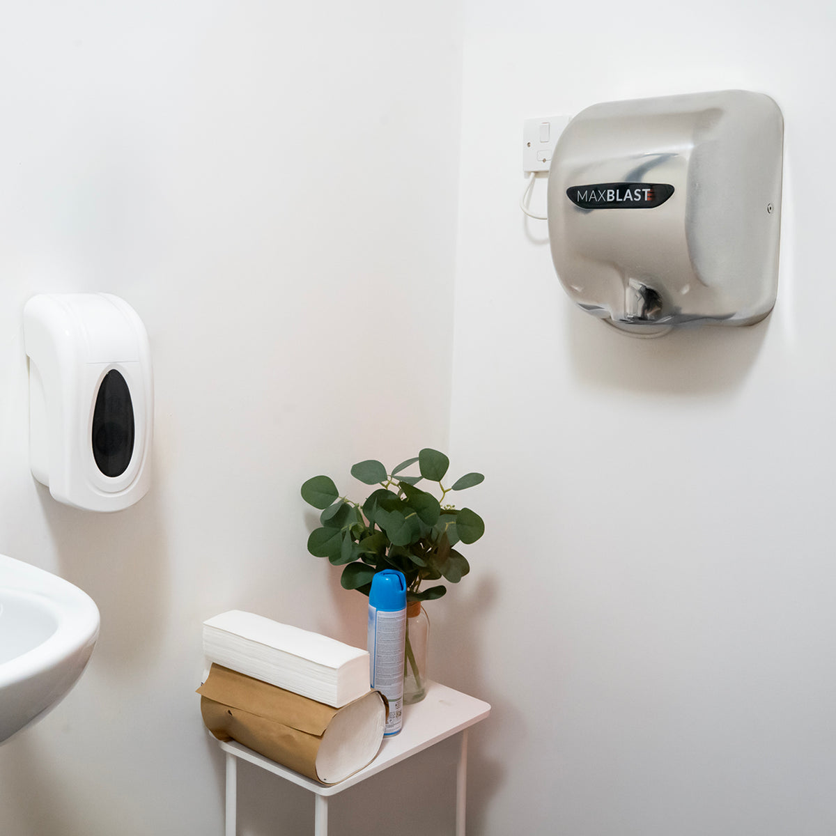 Secador de manos MAXBLAST en color plateado montado en la pared, junto a dispensador de jabón y planta decorativa en un baño moderno