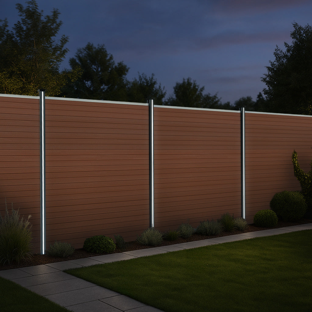 Valla exterior de WPC y aluminio en color marrón, con postes iluminados, rodeada de césped y plantas en un ambiente nocturno