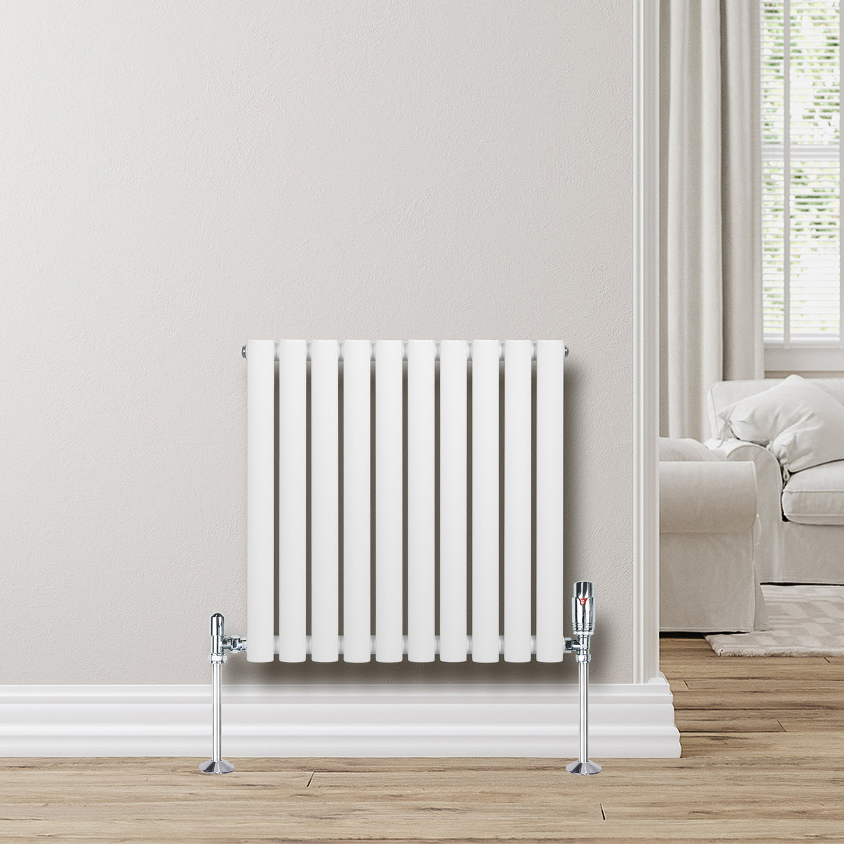 Radiador de columna ovalado blanco de 600 mm x 600 mm con válvulas, instalado en pared clara junto a un sofá y ventana iluminada