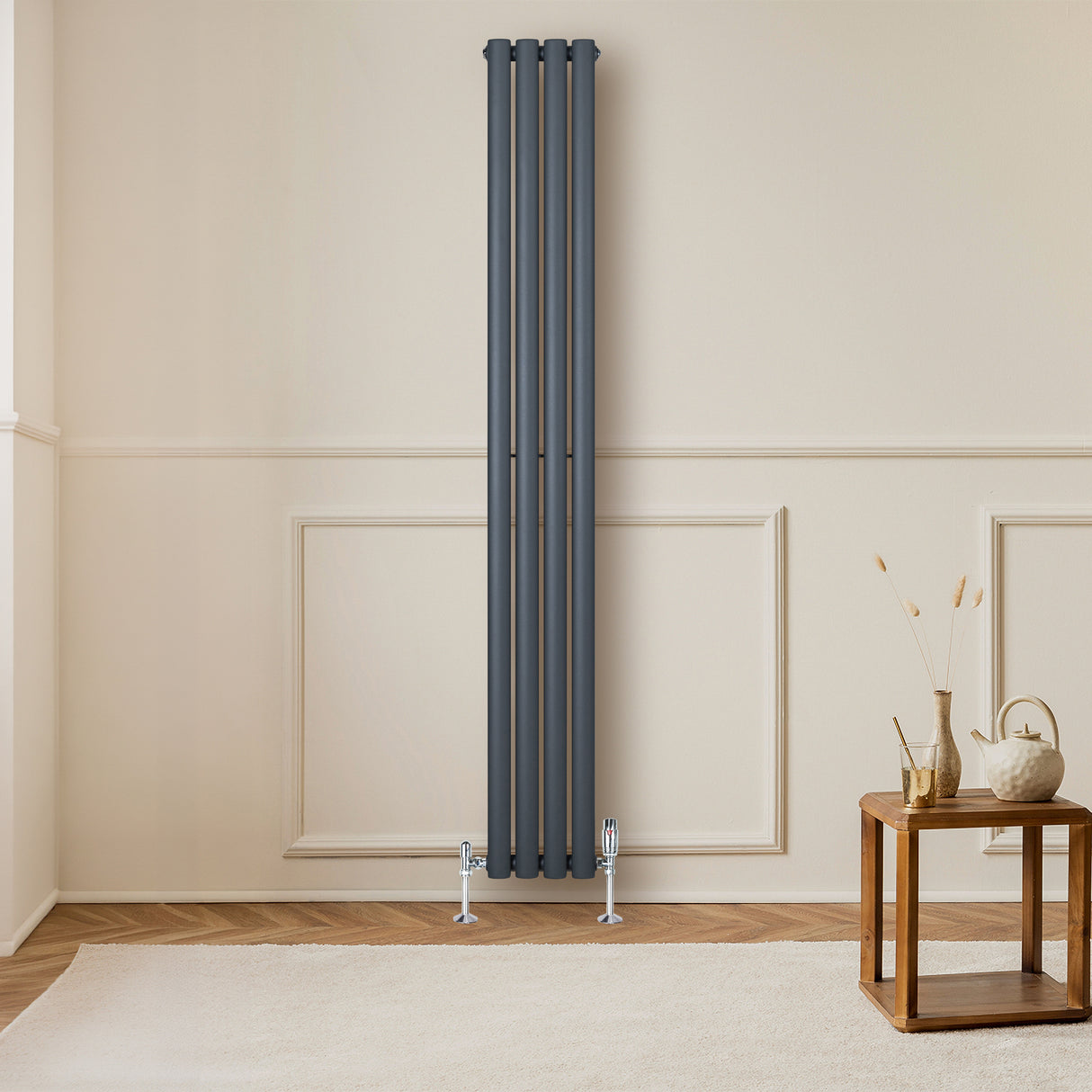 Radiador de columna ovalado en gris antracita, con válvulas, instalado en pared clara junto a una mesa de madera y decoración minimalista