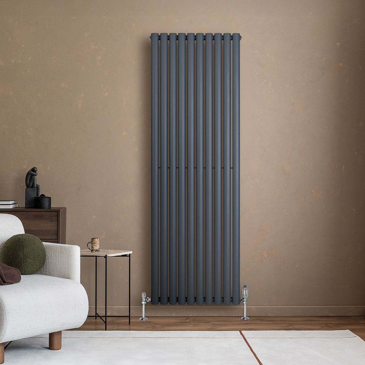 Radiador de columna ovalado en gris antracita, con diseño vertical y válvulas, instalado en pared de color claro, junto a un sofá moderno