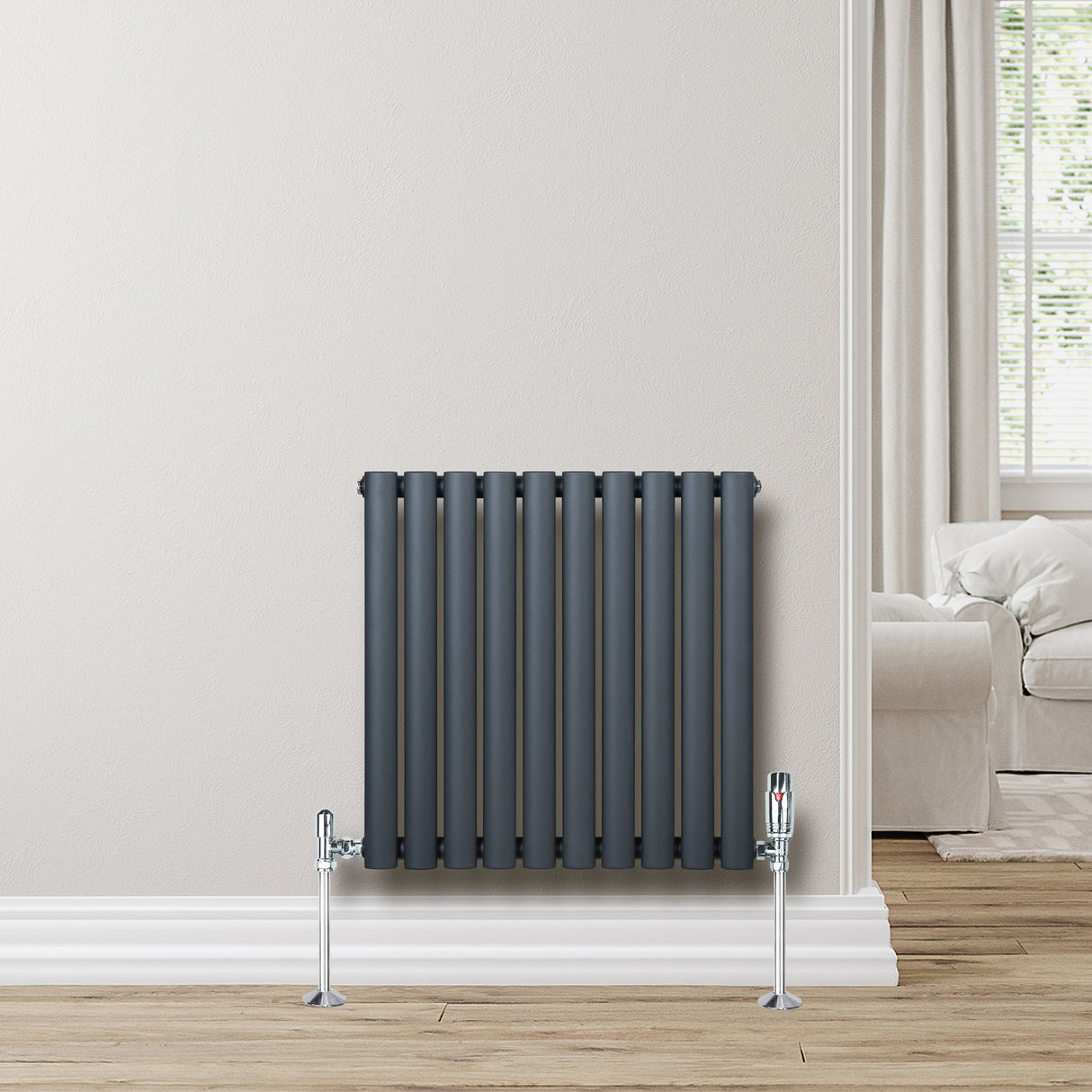 Radiador de columna ovalado en gris antracita, con válvulas metálicas, instalado en una pared clara junto a un sofá moderno