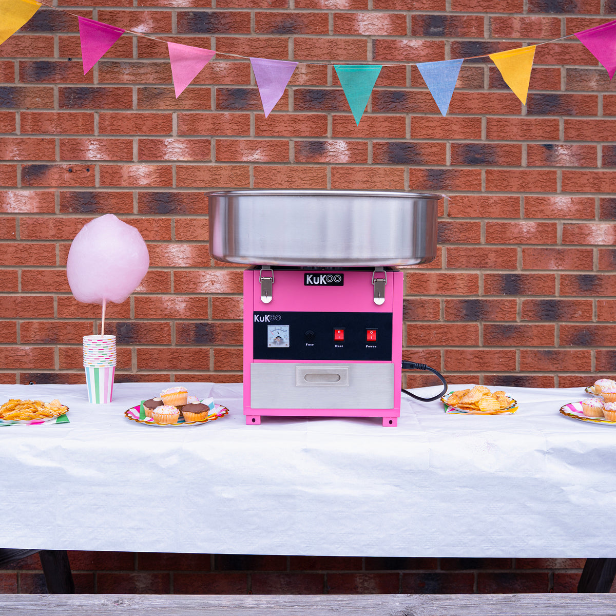 Máquina de algodón de azúcar KuKoo en color rosa, con tazón de acero inoxidable, decorada con banderines y golosinas en una mesa blanca