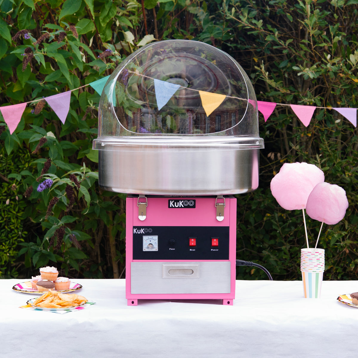 Máquina de algodón de azúcar KuKoo en color rosa con cúpula transparente, rodeada de decoraciones festivas y dulces en una mesa al aire libre