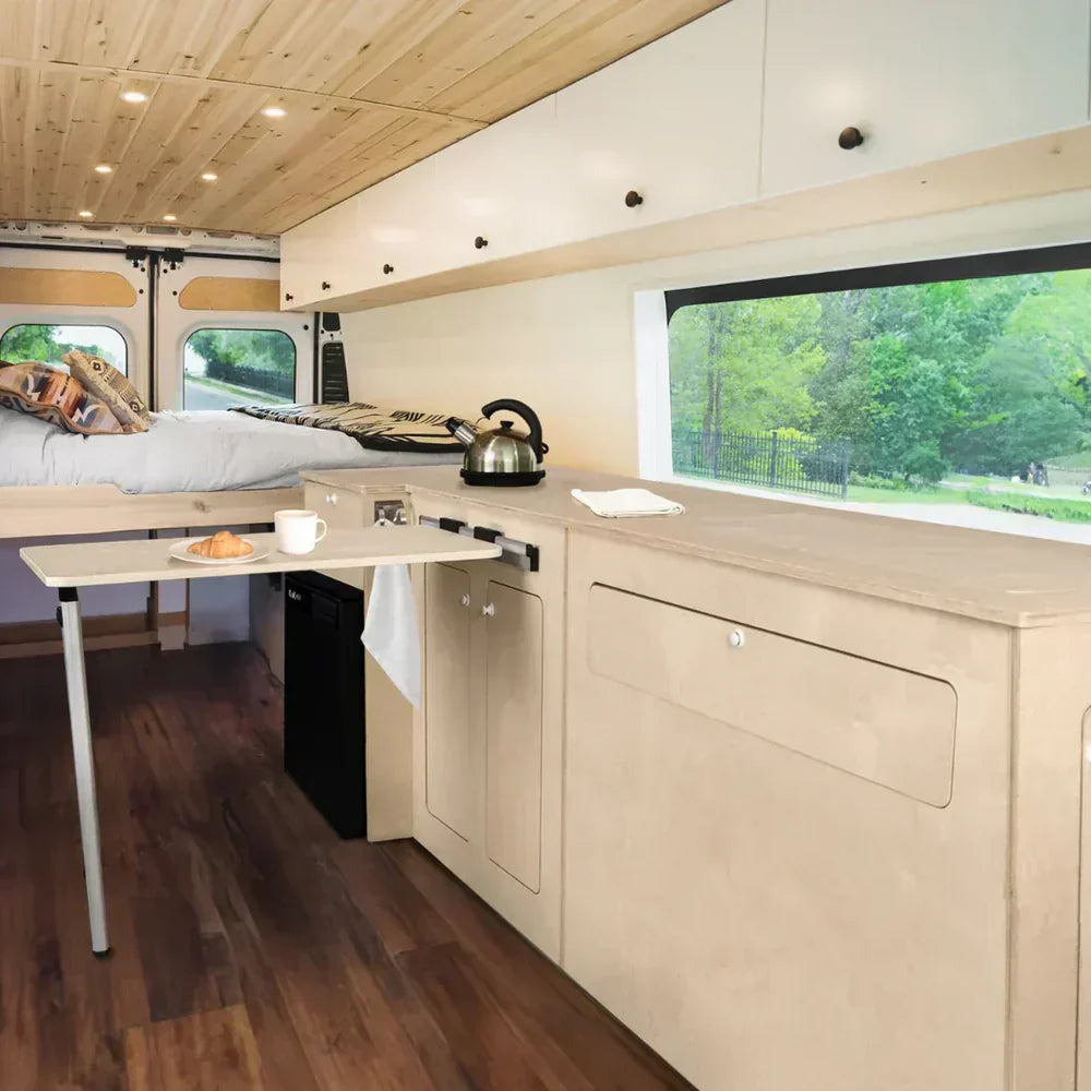 Interior de una furgoneta camper con muebles de madera clara, cama, cocina compacta y ventana que muestra un paisaje verde exterior