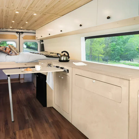 Interior de una furgoneta camper con muebles de madera clara, cama, cocina compacta y ventana que muestra un paisaje verde exterior