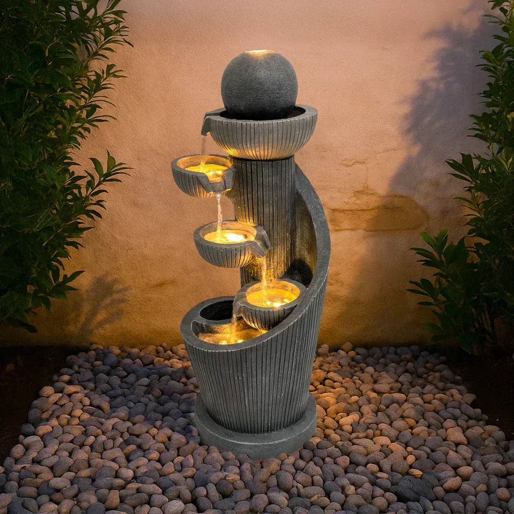 Fuente de agua eléctrica de piedra gris con luces, fluyendo en cascada sobre recipientes redondeados, rodeada de piedras decorativas