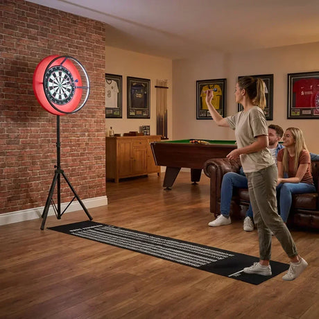 Mujer lanzando dardos hacia una diana iluminada, con amigos sentados en un sofá, en un ambiente acogedor de sala de juegos