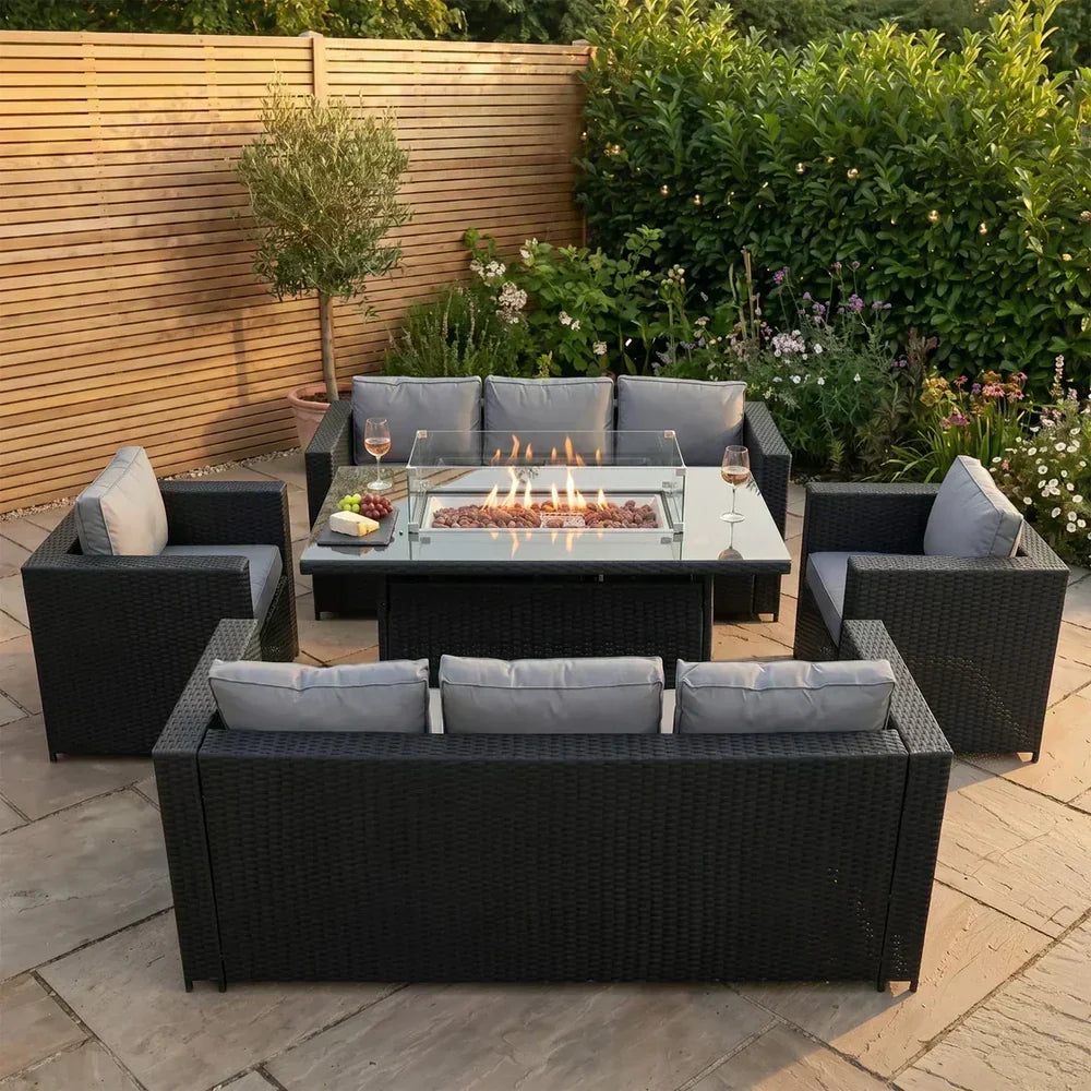 Conjunto de muebles de jardín de ratán negro con cojines grises, mesa de fuego en el centro, rodeado de plantas verdes y flores