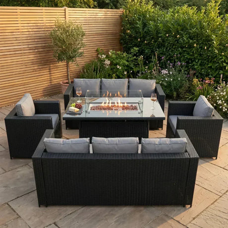 Conjunto de muebles de jardín de ratán negro con cojines grises, mesa de fuego en el centro, rodeado de plantas verdes y flores