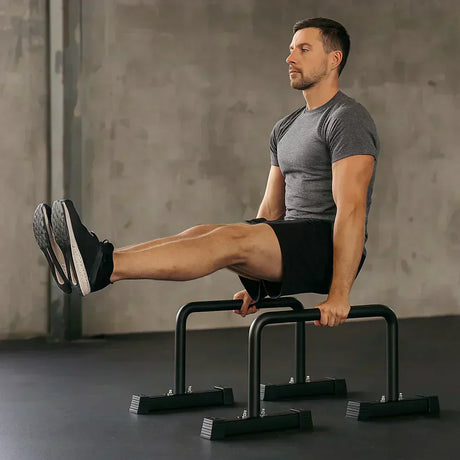 Hombre en camiseta gris y pantalones cortos realizando ejercicios de calistenia en paralelas negras sobre un suelo oscuro