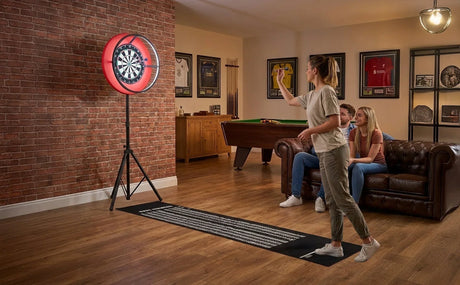 Diana autoportante roja en una sala de juegos, mujer lanzando dardos, sofá marrón y amigos observando en un ambiente acogedor