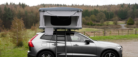 Tienda de techo gris montada sobre un SUV plateado, rodeada de un paisaje natural con árboles y un camino de tierra