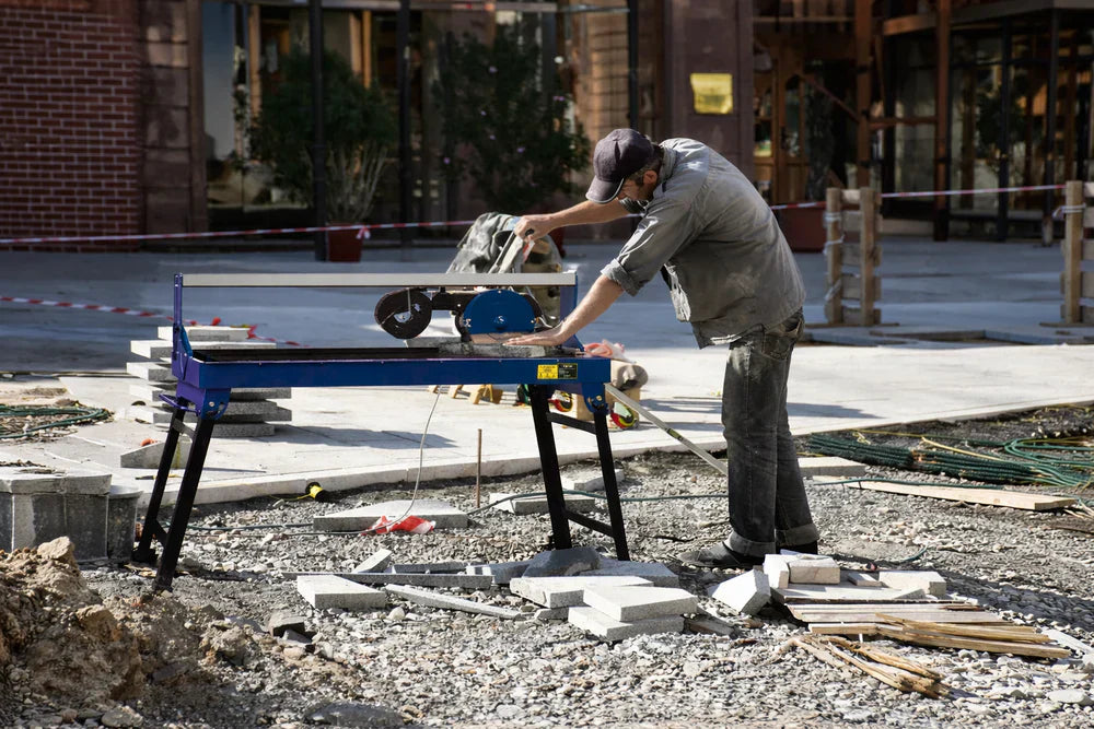 Hombre trabajando en una cortadora de azulejos húmeda, rodeado de materiales de construcción y un entorno de obra al aire libre