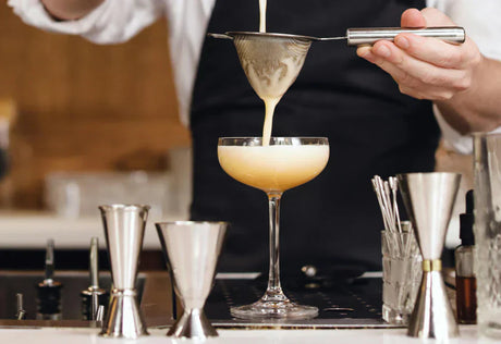 Coctel amarillo vertido en una copa elegante, con un colador de metal, rodeado de herramientas de coctelería brillantes y pulidas