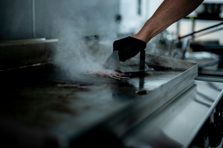 Superficie de una plancha grill eléctrica con vapor, mientras una mano con guante negro voltea carne dorada con espátula metálica