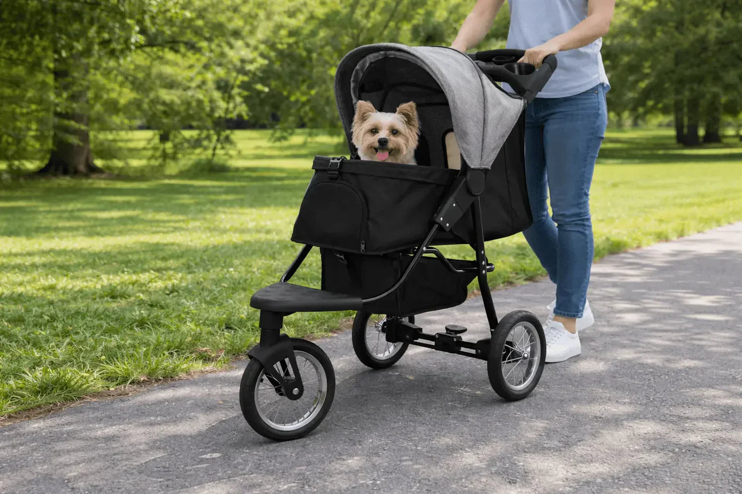 Carrito para mascotas negro con detalles grises, paseando por un parque, con un perro pequeño feliz en su interior