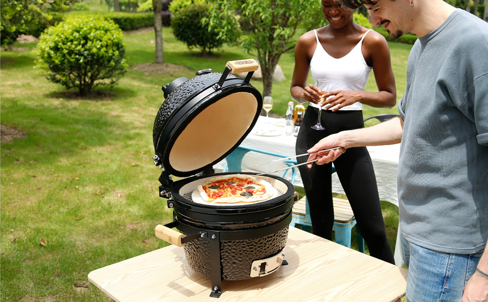 Parrilla Kamado negra con pizza colorida en su interior, rodeada de personas disfrutando al aire libre en un jardín verde