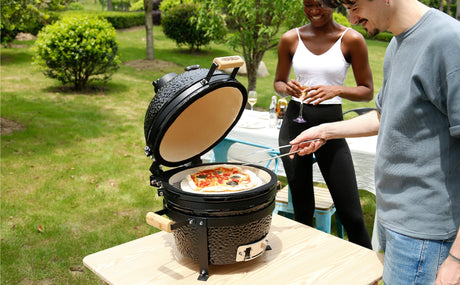 Parrilla Kamado negra con pizza colorida en su interior, rodeada de personas disfrutando al aire libre en un jardín verde