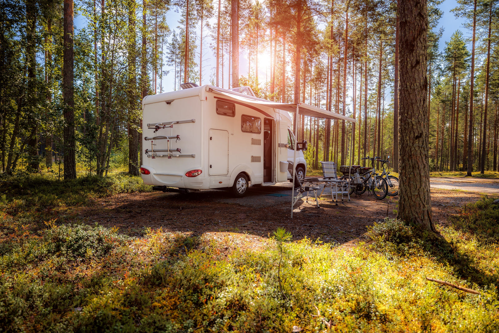 Furgoneta blanca estacionada en un bosque, con sillas plegables y bicicletas, rodeada de árboles y luz solar filtrándose entre las hojas