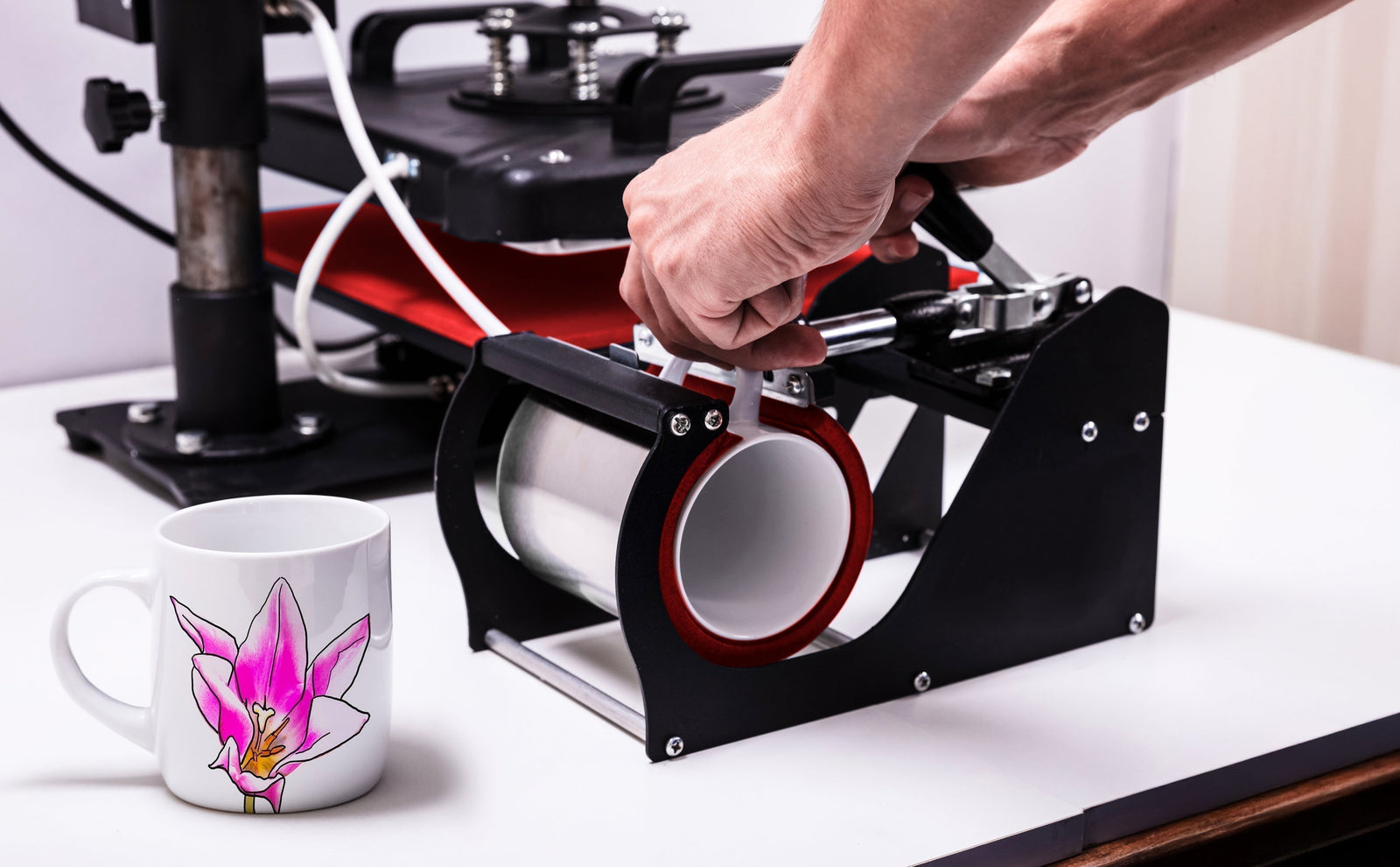 Prensa térmica para tazas con diseño negro y rojo, mano ajustando el rodillo, taza blanca decorada con una flor rosa al lado