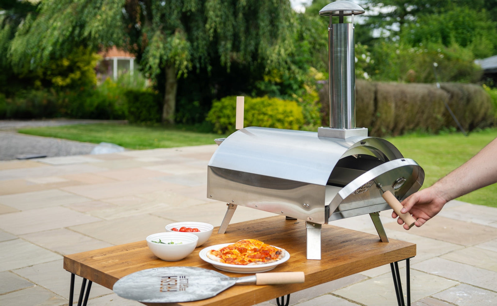 Horno de pizza de acero inoxidable en un jardín, con pizza recién horneada y ingredientes frescos en platos sobre una mesa de madera