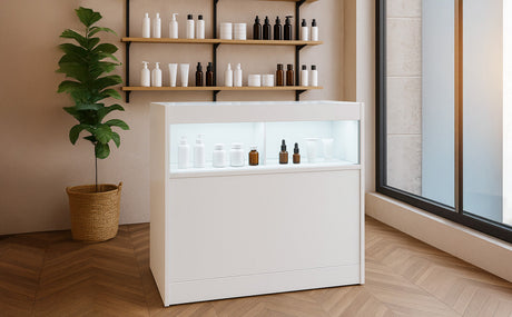 Mostrador blanco moderno con iluminación, exhibiendo productos de belleza en frascos variados, rodeado de estanterías y plantas decorativas