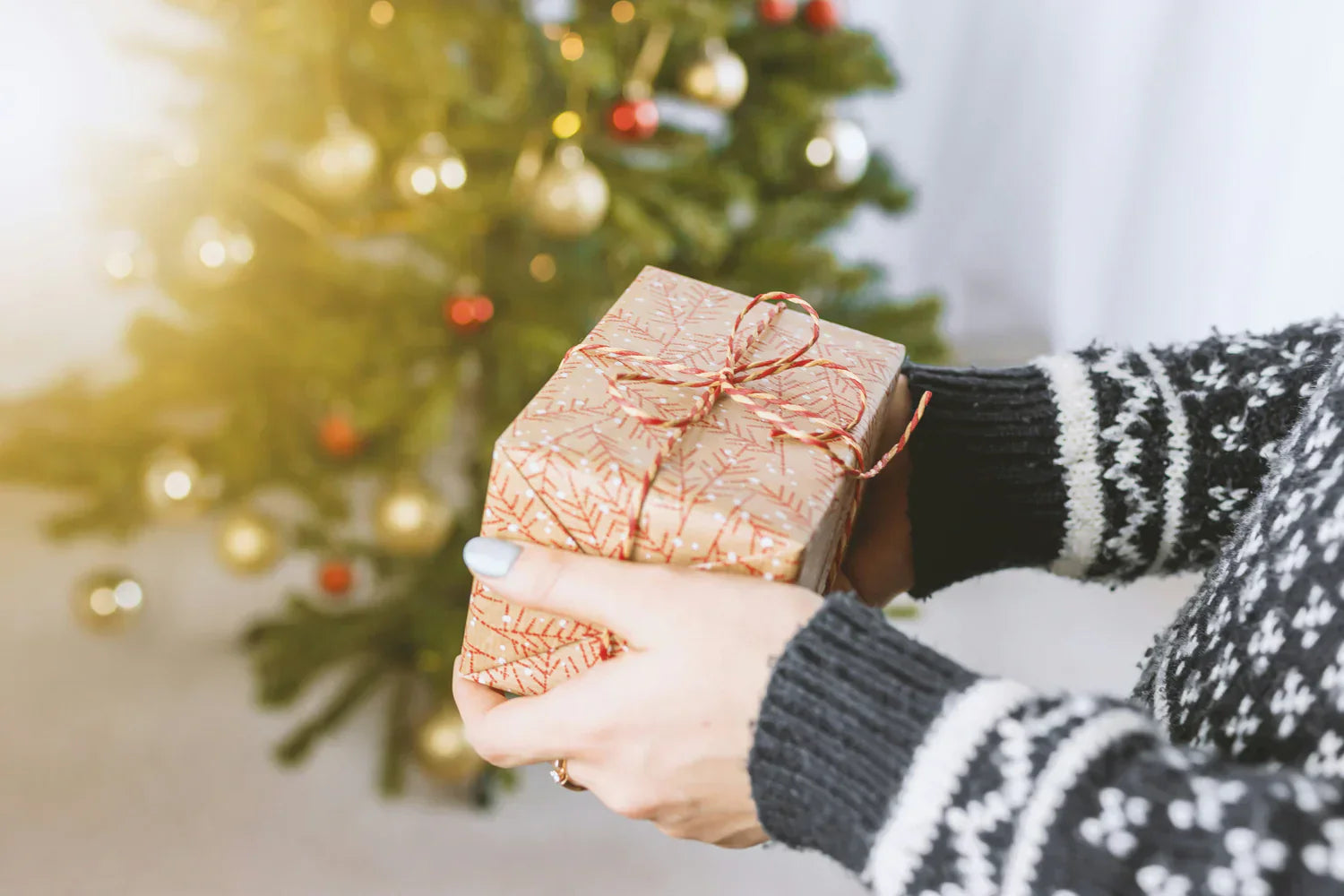 Regalo envuelto en papel dorado con diseño de copos de nieve, sostenido por manos con suéter negro, frente a un árbol de Navidad decorado