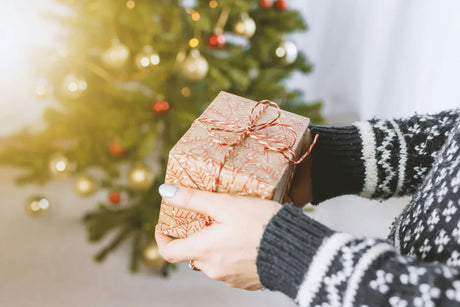 Regalo envuelto en papel dorado con diseño de copos de nieve, sostenido por manos con suéter negro, frente a un árbol de Navidad decorado