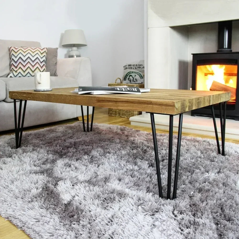 Mesa de café de madera clara con patas de horquilla negras, sobre una alfombra suave y gris, junto a una chimenea encendida