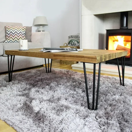 Mesa de café de madera clara con patas de horquilla negras, sobre una alfombra suave y gris, junto a una chimenea encendida
