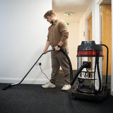 Aspiradora comercial MAXBLAST de 80 litros, con diseño robusto, manguera flexible y usuario limpiando una alfombra gris en un pasillo
