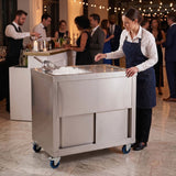 Carrito de bar de acero inoxidable con hielo, una bartender lo utiliza en un evento elegante con invitados de fondo y luces brillantes