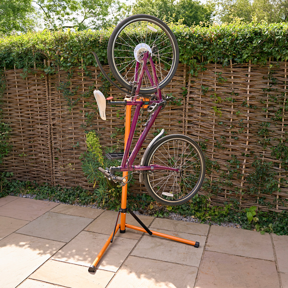 Soporte de reparación para bicicletas en color naranja y negro, sostiene una bicicleta de montaña de color púrpura al revés sobre un fondo de seto verde