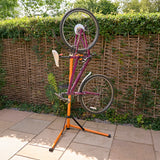 Soporte de reparación para bicicletas en color naranja y negro, sostiene una bicicleta de montaña de color púrpura al revés sobre un fondo de seto verde
