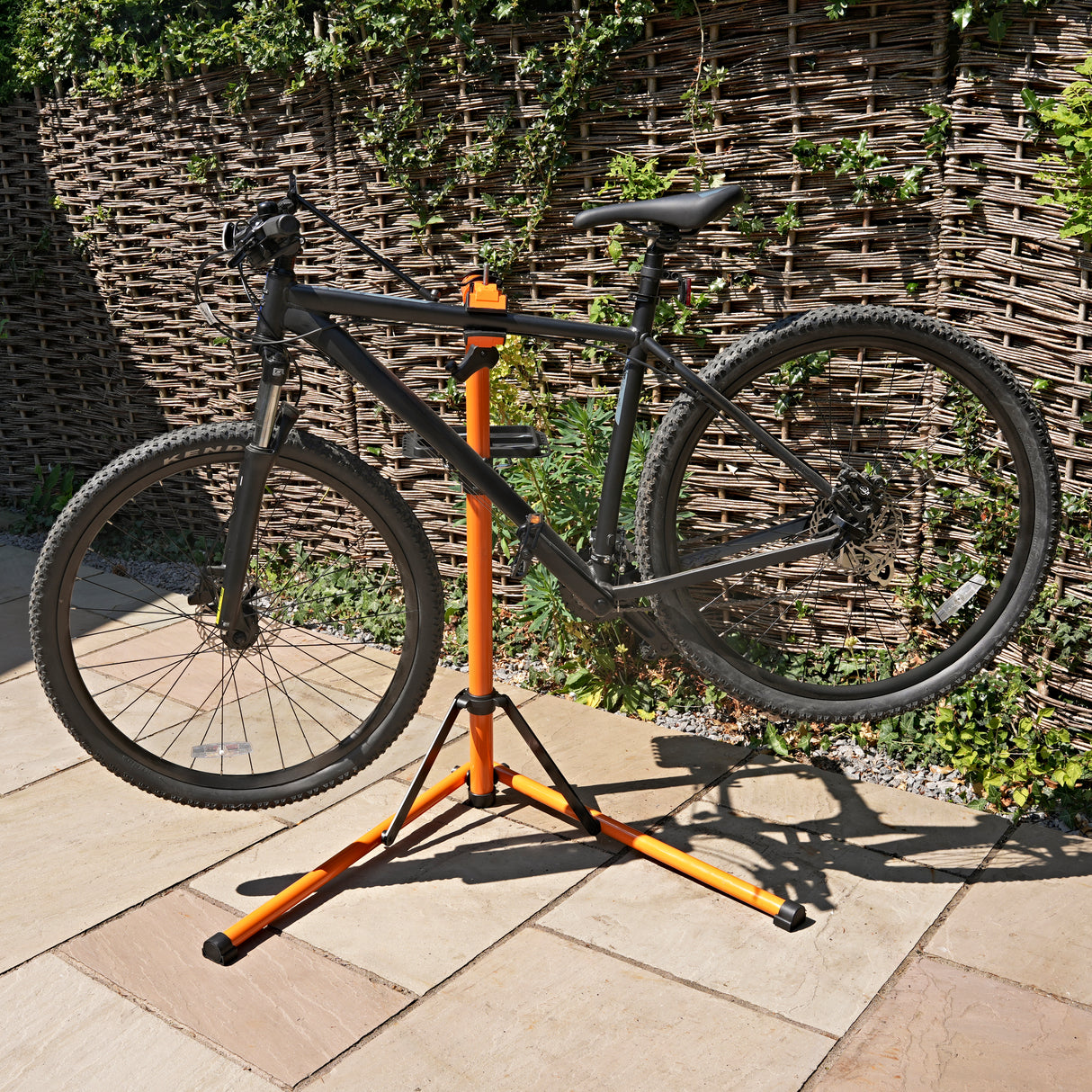 Soporte de reparación para bicicletas en color negro y naranja, con un diseño robusto y ajustable, sosteniendo una bicicleta de montaña