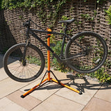 Soporte de reparación para bicicletas en color negro y naranja, con un diseño robusto y ajustable, sosteniendo una bicicleta de montaña