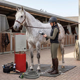 Ducha portátil roja para caballos, mujer usando esquiladora inalámbrica, caballo gris en establo, ambiente limpio y organizado