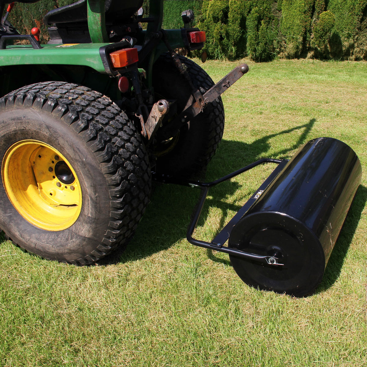 Rodillo de jardín remolcable T-Mech de 120 L, cilindro negro brillante con marco metálico, acoplado a un tractor en césped verde