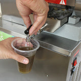 Máquina exprimidora de caña de azúcar de acero inoxidable, con un vaso transparente lleno de jugo marrón claro y una mano sujetando el vaso