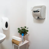 Secador de manos MAXBLAST en color plateado montado en la pared, junto a dispensador de jabón y planta decorativa en un baño moderno