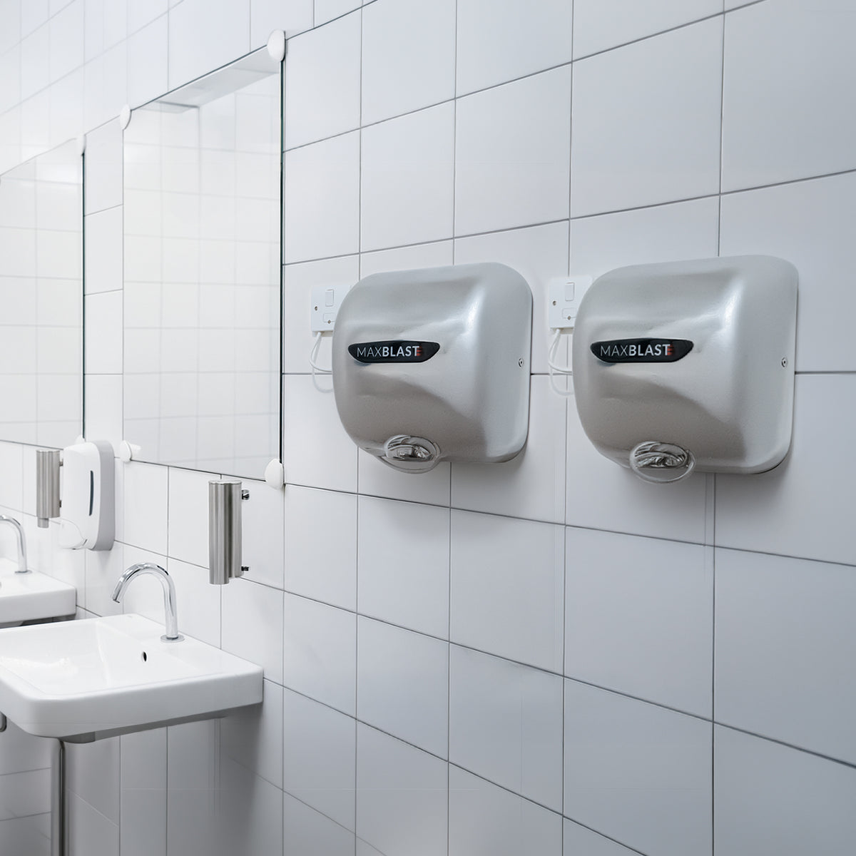 Secadores de manos comerciales MAXBLAST en acabado plateado, instalados en una pared blanca de azulejos, junto a un lavabo moderno