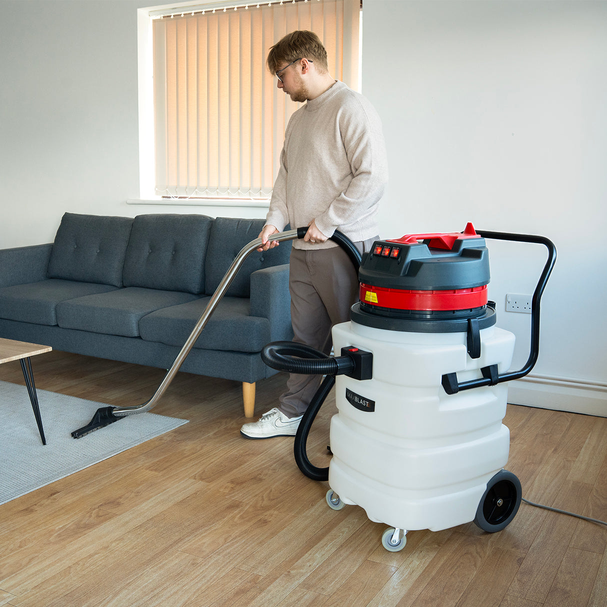Aspirador industrial de sólidos y líquidos de 90 L, con manguera y boquilla, en un ambiente moderno con sofá gris y suelo de madera clara