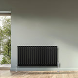 Radiador de columna ovalado negro de 600 mm x 1200 mm, con válvulas, instalado en una pared junto a una ventana con vista al exterior