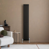 Radiador de columna ovalado negro de 1600 mm x 240 mm, con válvulas, instalado en pared beige junto a un sofá claro y mesa de madera