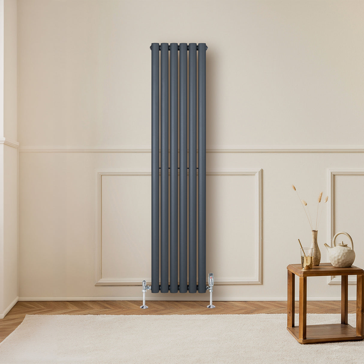 Radiador de columna ovalado en gris antracita, con válvulas, instalado en pared clara, junto a una mesa de madera y decoración minimalista