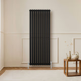 Radiador de columna ovalado negro de 1600 mm x 600 mm, con válvulas, instalado en una pared clara junto a una mesa de madera