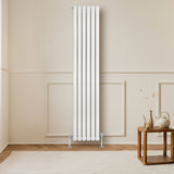 Radiador de columna ovalado en blanco, con diseño vertical y válvulas, instalado en una pared clara, junto a una mesa de madera