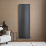 Radiador de columna ovalado en gris antracita, con diseño vertical y válvulas, instalado en pared de color claro, junto a un sofá moderno